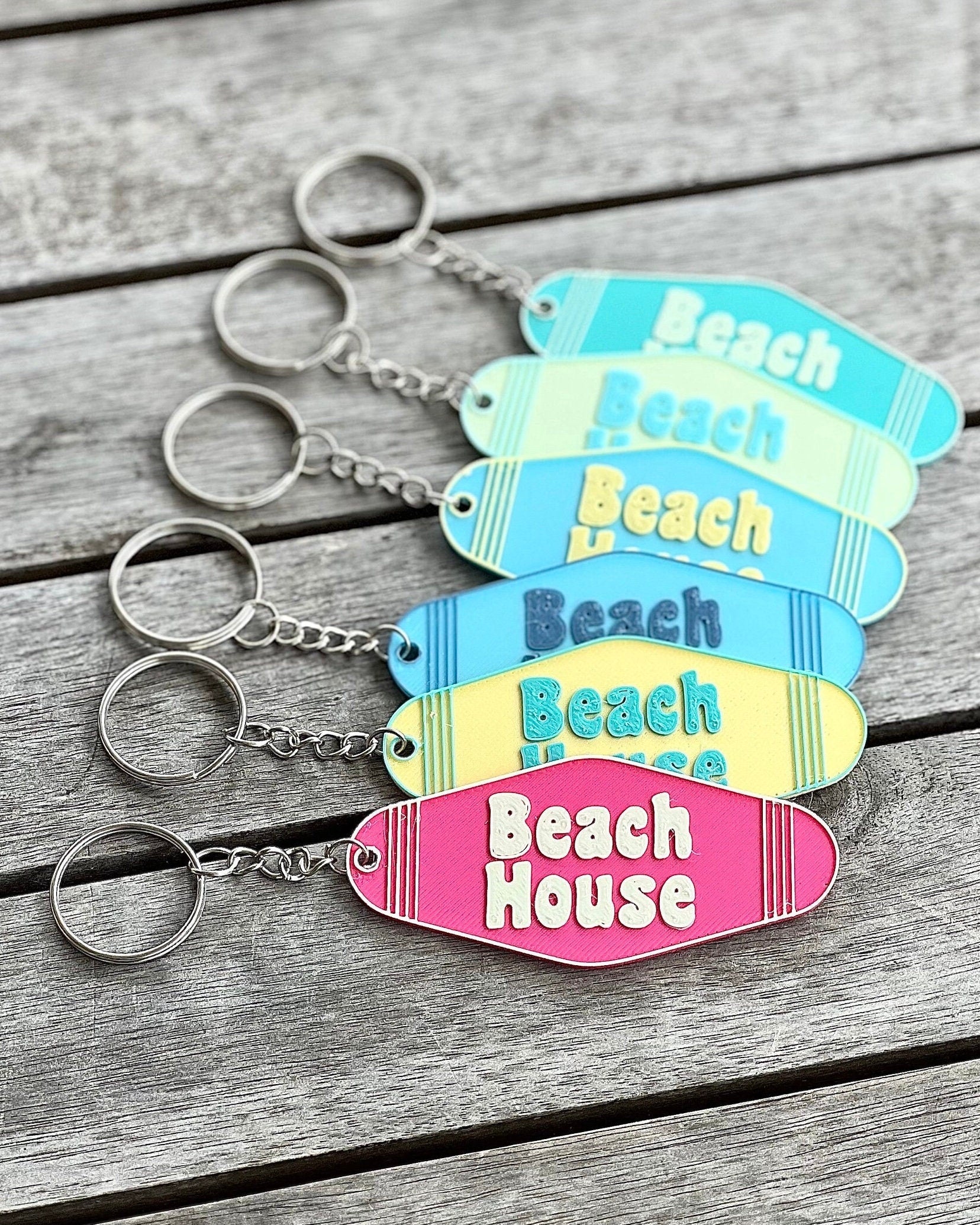Five 3D printed Beach House motel-style keychains in blue, teal, yellow, and pink with key rings on a wood surface
