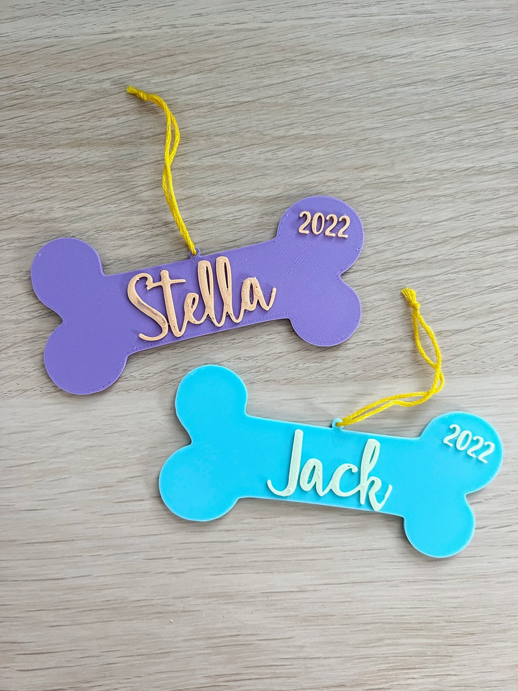 Purple Stella and blue Jack 3D printed dog bone ornaments with yellow hanging string on a wood surface
