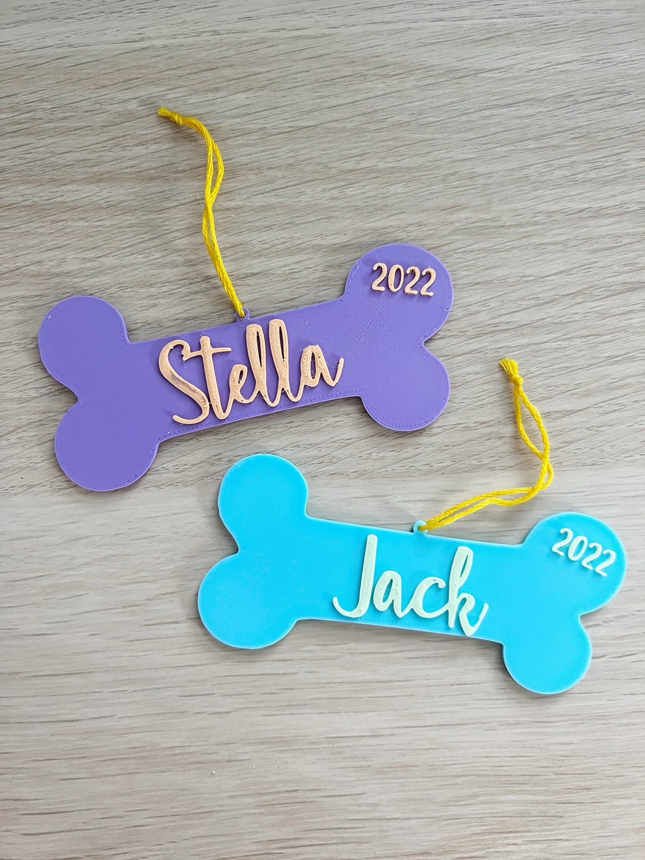 Purple Stella and blue Jack 3D printed dog bone ornaments with yellow hanging string on a wood surface