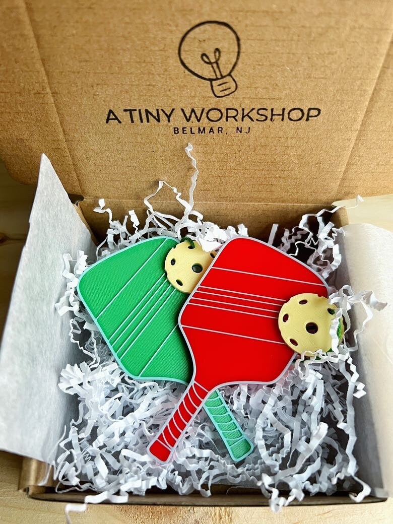 Green and red pickleball paddle ornaments packed in an A Tiny Workshop gift box with white crinkle paper