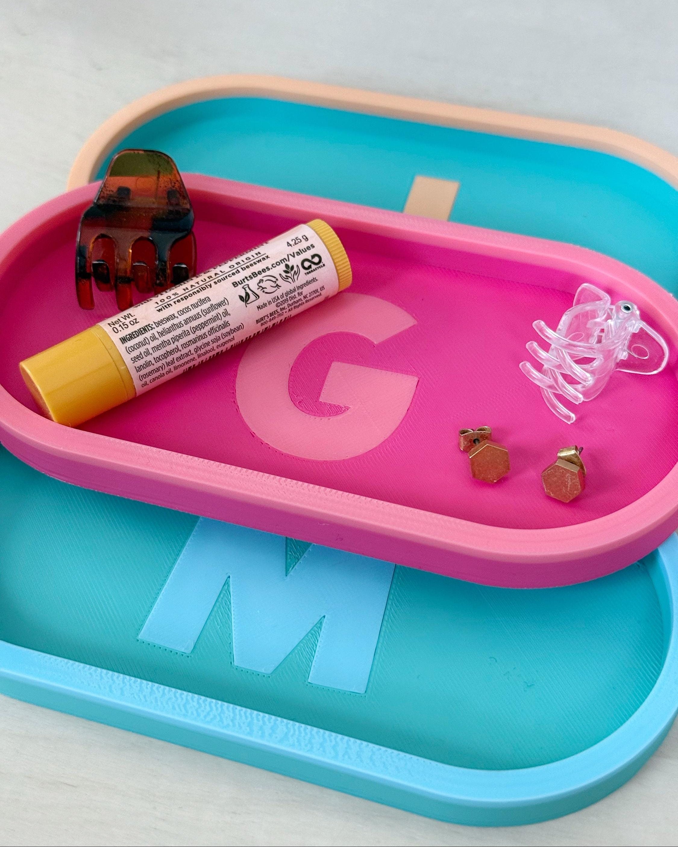 Three personalized initial catch-all trays in pink, teal, and blue holding a lip balm, hair clip, and earrings