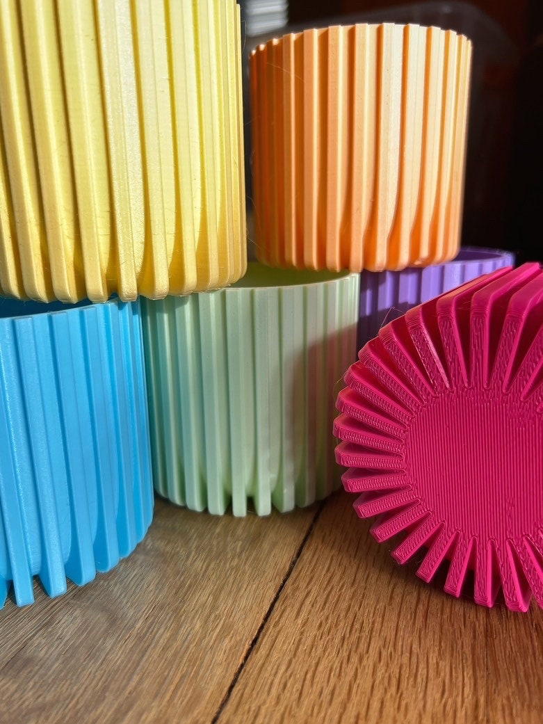 Stack of 3D printed Modern Fluted Planters in yellow, peach, light blue, mint green and lavender next to a magenta fluted pot on a wood table