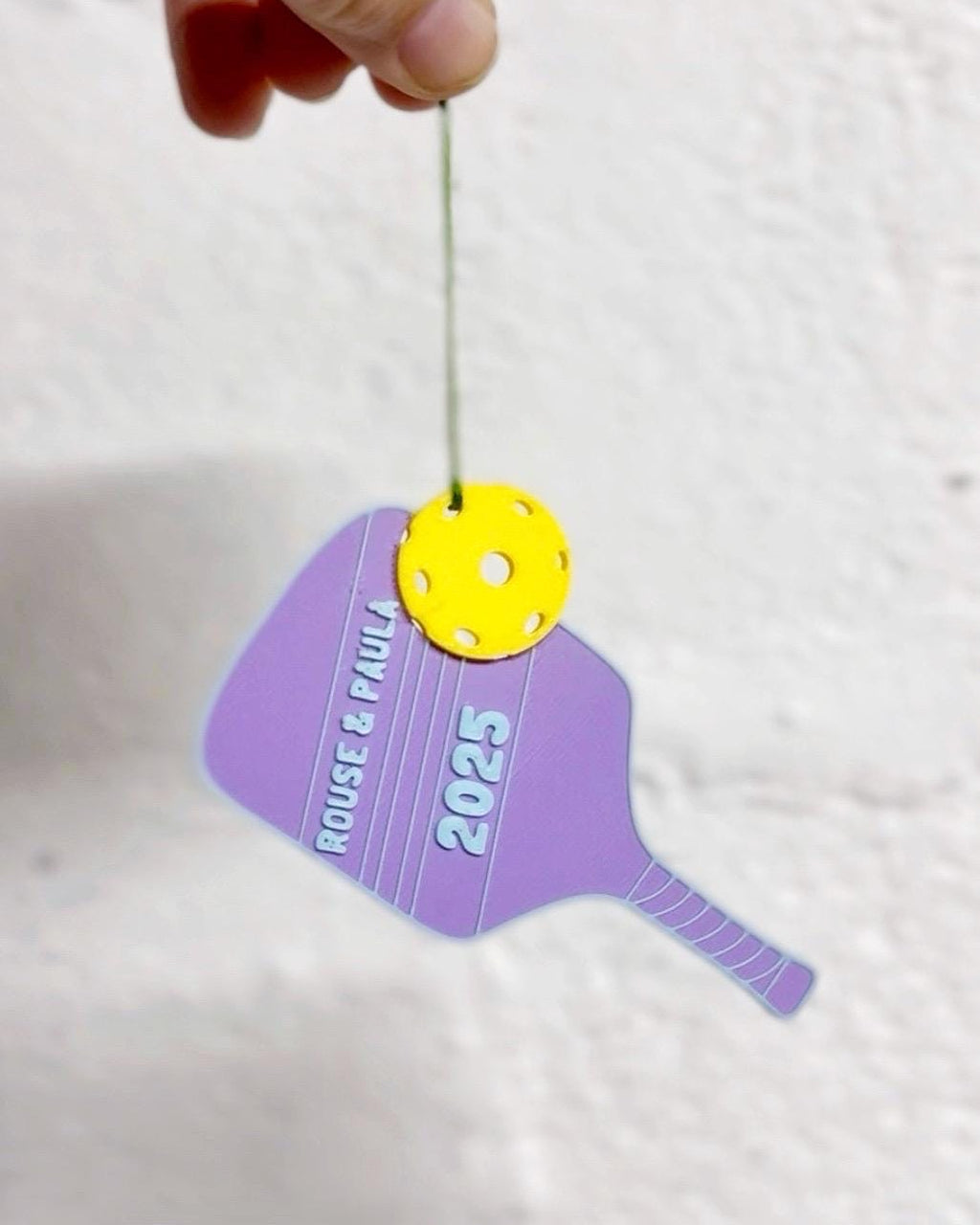 Personalized purple pickleball paddle ornament reading Rouse and Paul 2025 with a yellow ball, held by hand