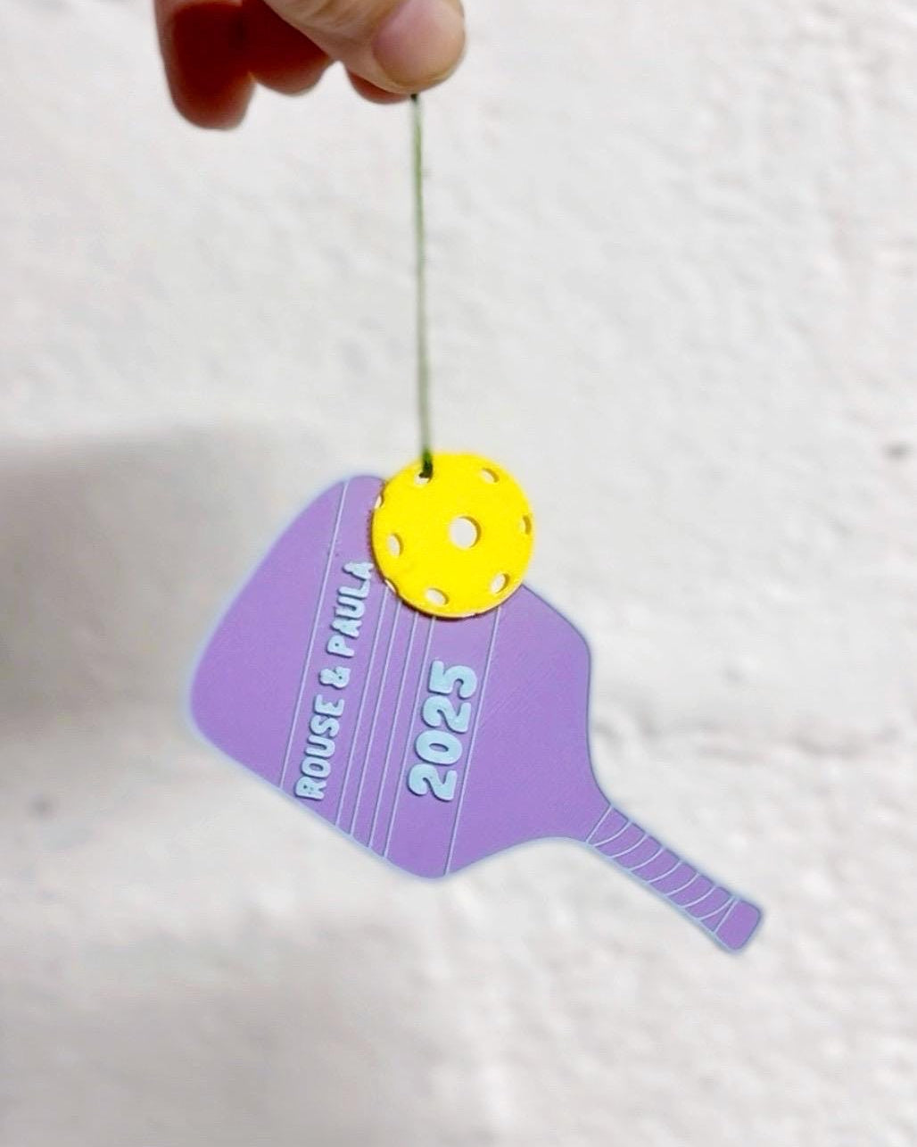 Personalized purple pickleball paddle ornament reading Rouse and Paul 2025 with a yellow ball, held by hand