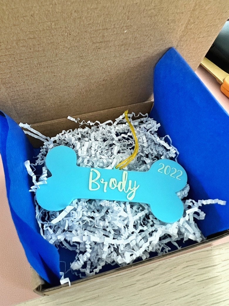 Personalized dog bone ornament reading Brody packed in a gift box with blue tissue and white crinkle paper