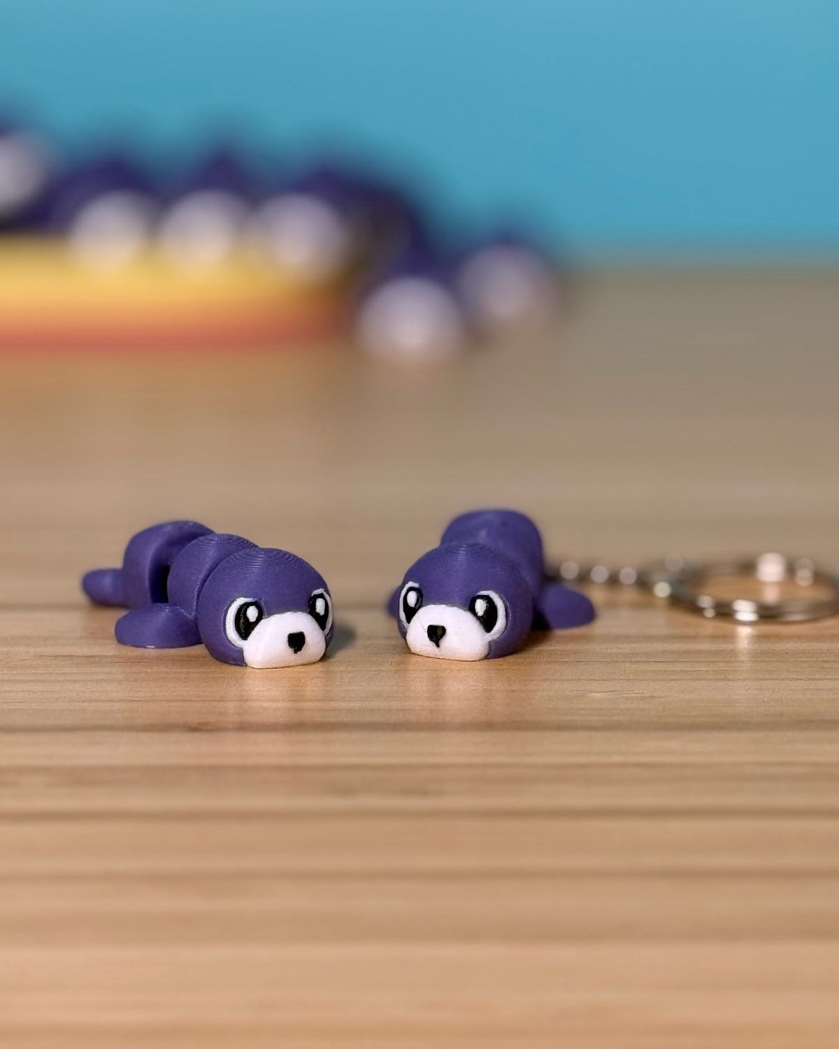 Two purple 3D printed Mini Flexi Seal fidget toys on a wood desk, one with a keychain ring