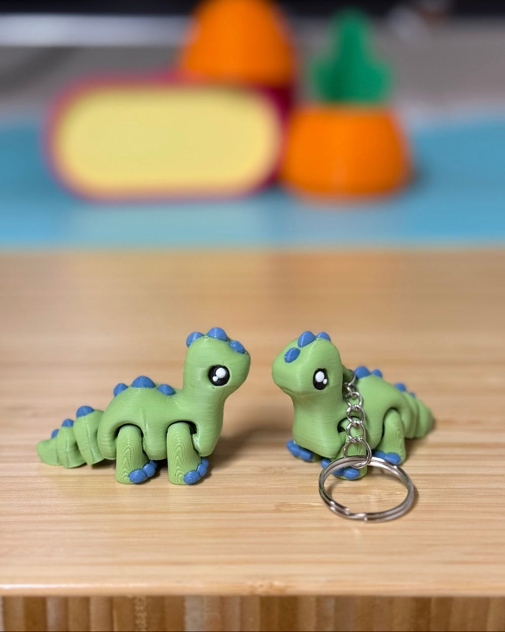 Two green 3D printed Mini Flexi Brontosaurus fidget toys on a wood desk, one with a keychain ring