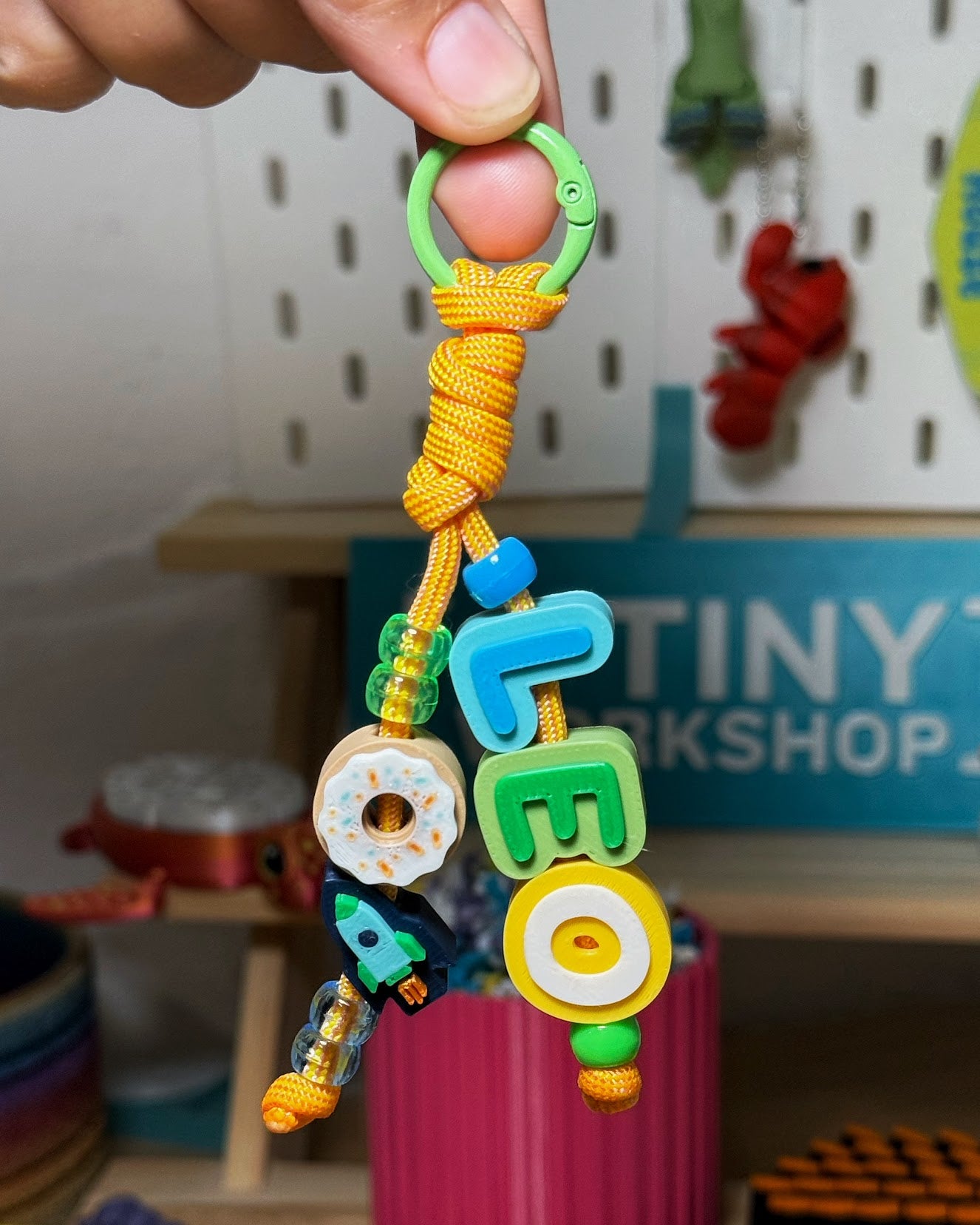 Hand holding a 3D printed Beaded Name Keychain spelling LEO with donut and rocket charms in front of the A Tiny Workshop pegboard