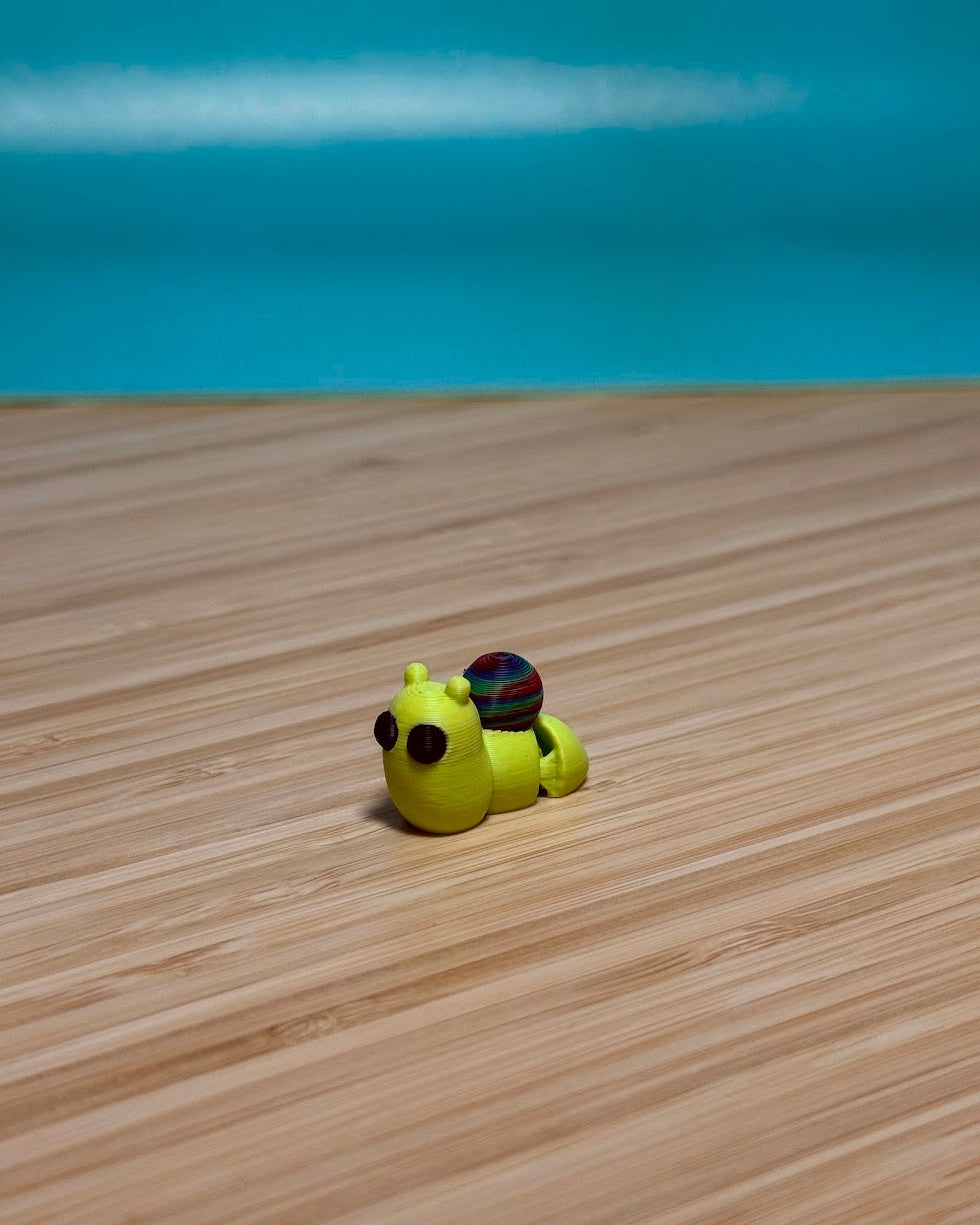 Yellow 3D printed Mini Flexi Snail fidget toy with a purple shell on a wood desk