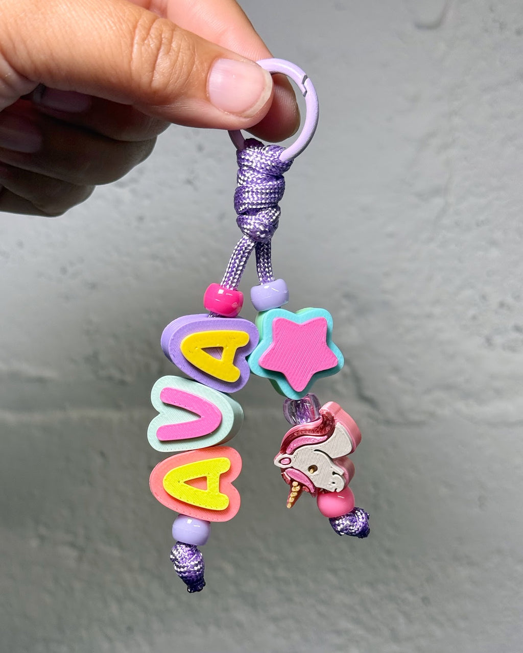 Hand holding a 3D printed Beaded Name Keychain spelling AVA in pink, yellow and blue letter beads with a star and a unicorn charm on purple cord
