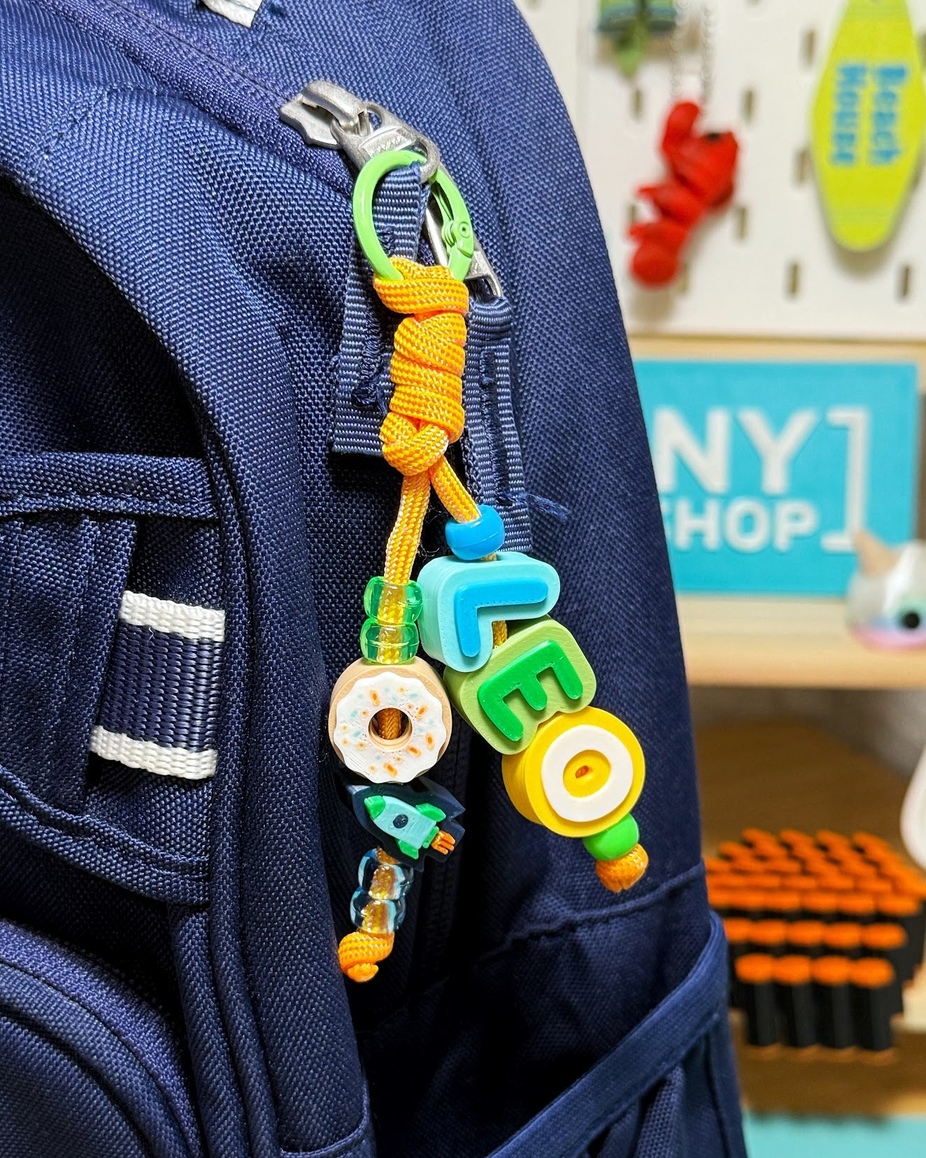Beaded Name Keychain spelling LEO on yellow cord with a donut and a rocket charm, clipped to a navy backpack