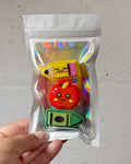 A hand holding a clear, iridescent zip-top pouch containing three cute, cartoon-style erasers shaped like a yellow pencil, a red apple, and a green crayon, against a light gray textured wall.