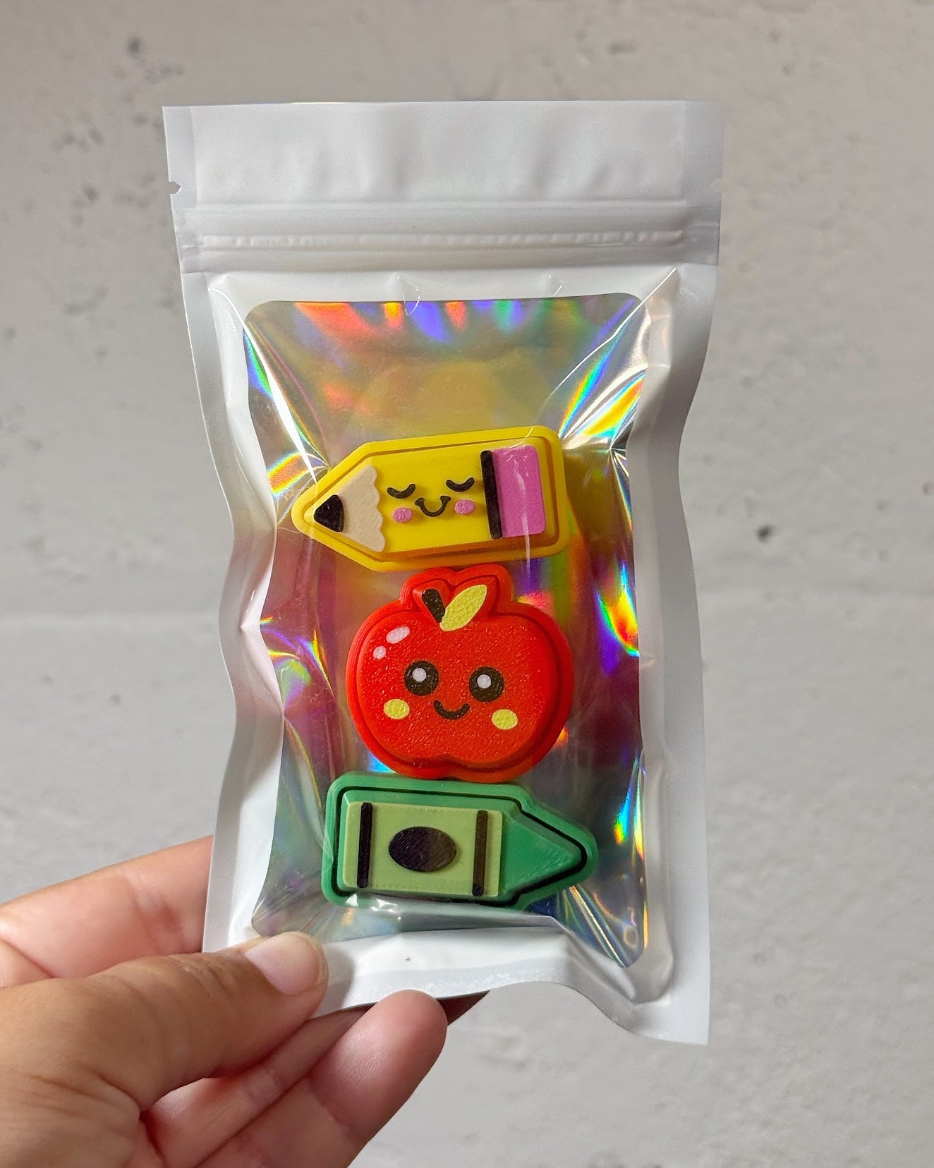 A hand holding a clear, iridescent zip-top pouch containing three cute, cartoon-style erasers shaped like a yellow pencil, a red apple, and a green crayon, against a light gray textured wall.