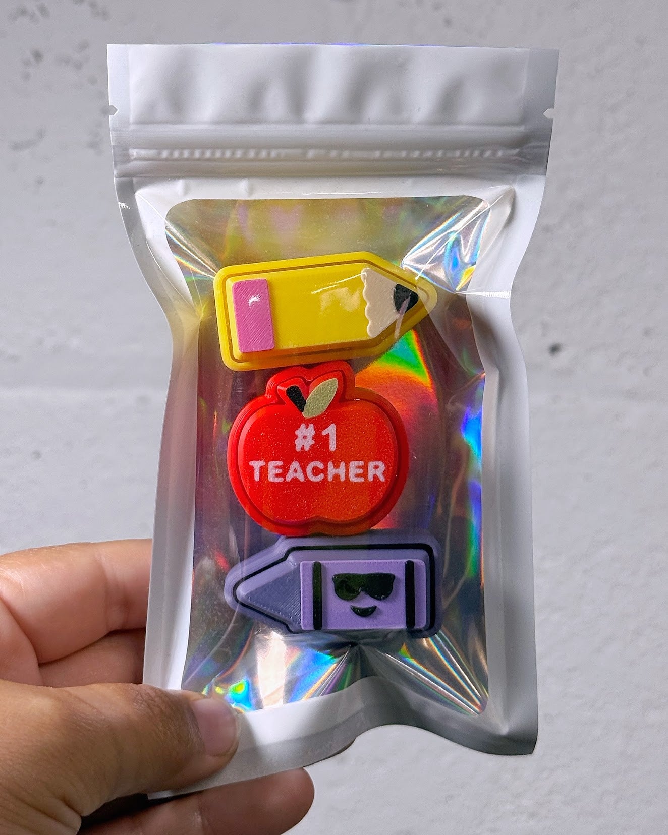 Teacher Clicky Pack pouch with a pencil, a red apple reading Number 1 Teacher, and a purple crayon clicky, held in a hand