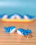 Two blue 3D printed Mini Flexi Whale fidget toys on a wood desk, one with a keychain ring