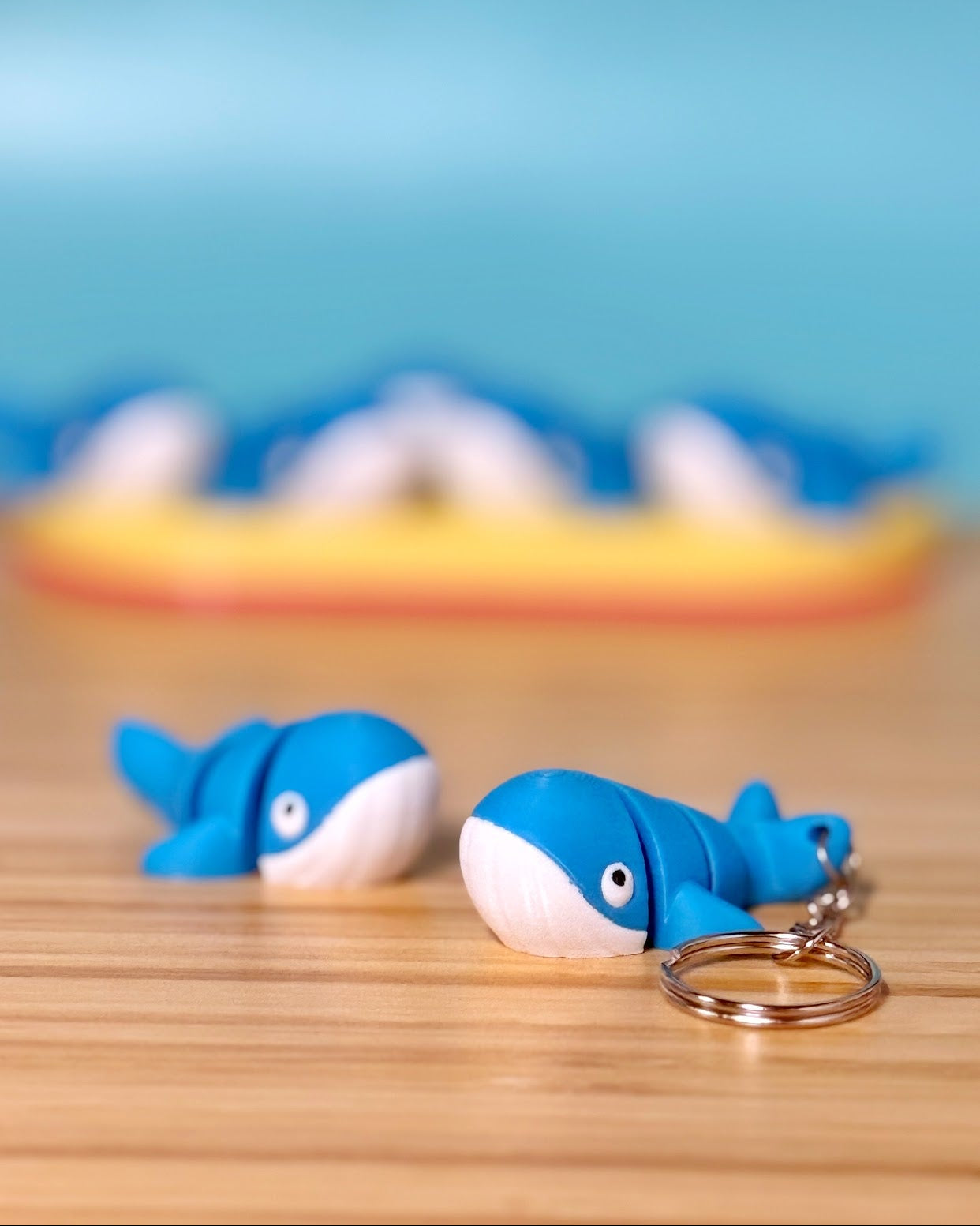 Two blue 3D printed Mini Flexi Whale fidget toys on a wood desk, one with a keychain ring