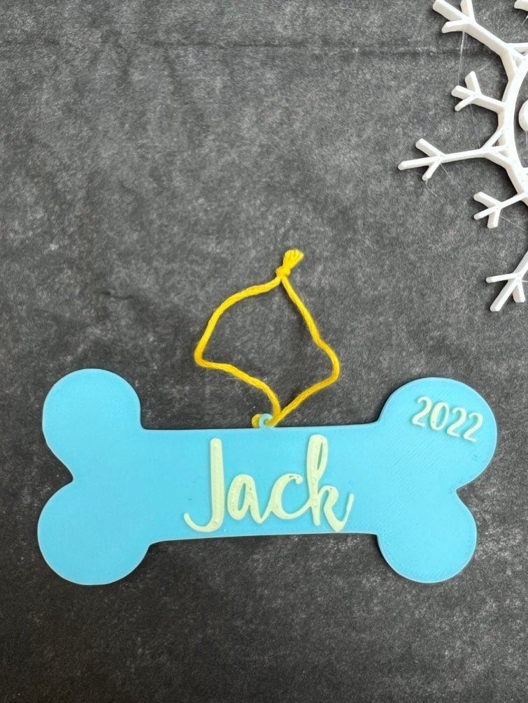 Blue 3D printed dog bone ornament reading Jack 2022 with yellow string on a dark background