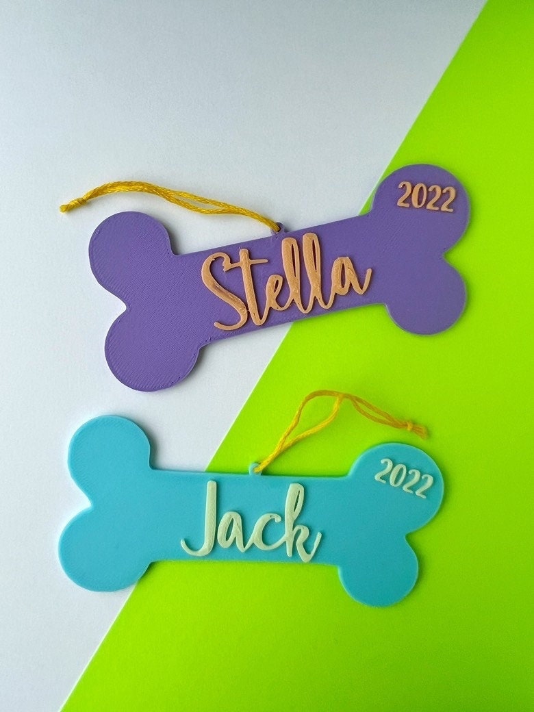 Two personalized dog bone ornaments, purple with peach lettering reading Stella and blue with mint reading Jack, on a green and white background