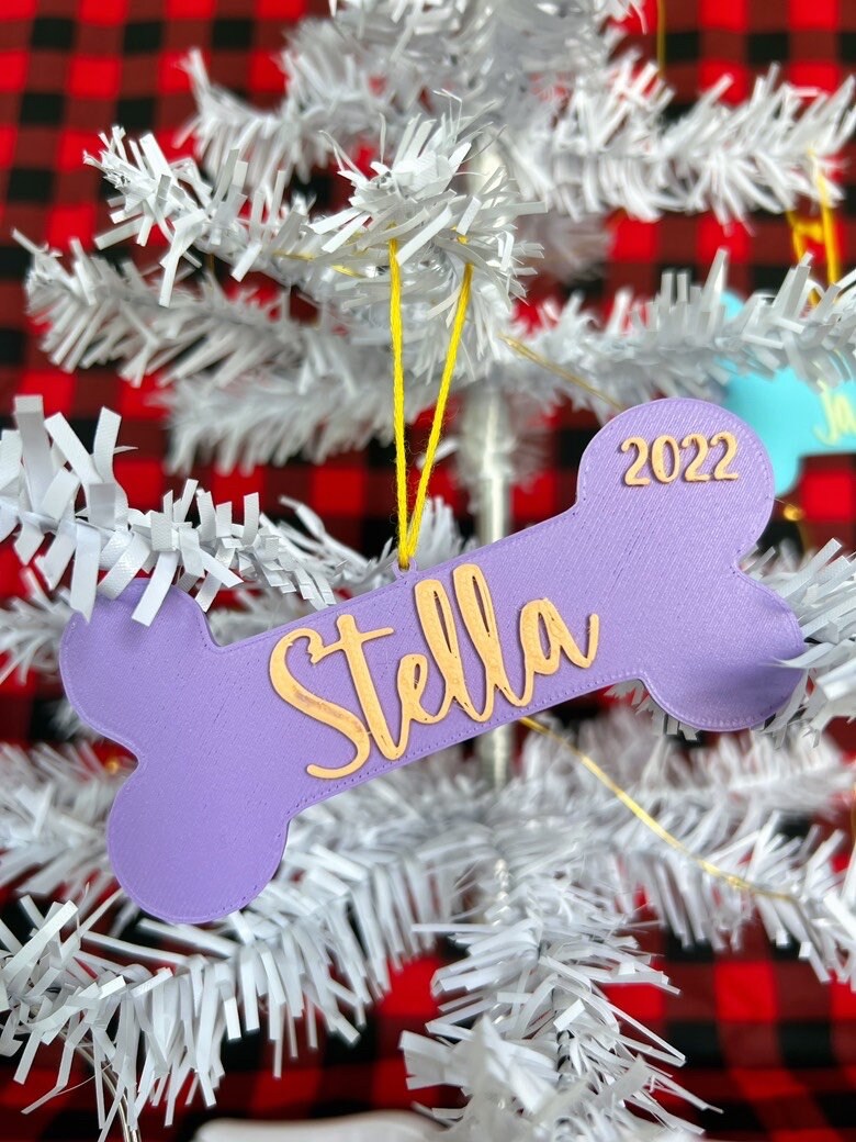 Purple personalized dog bone ornament reading Stella hanging on a white Christmas tree
