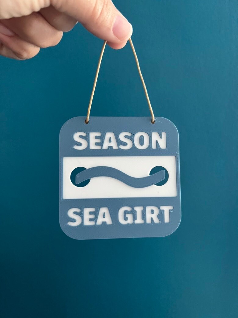 Hand holding a 3D printed Sea Girt beach badge ornament on a twine loop against a teal wall