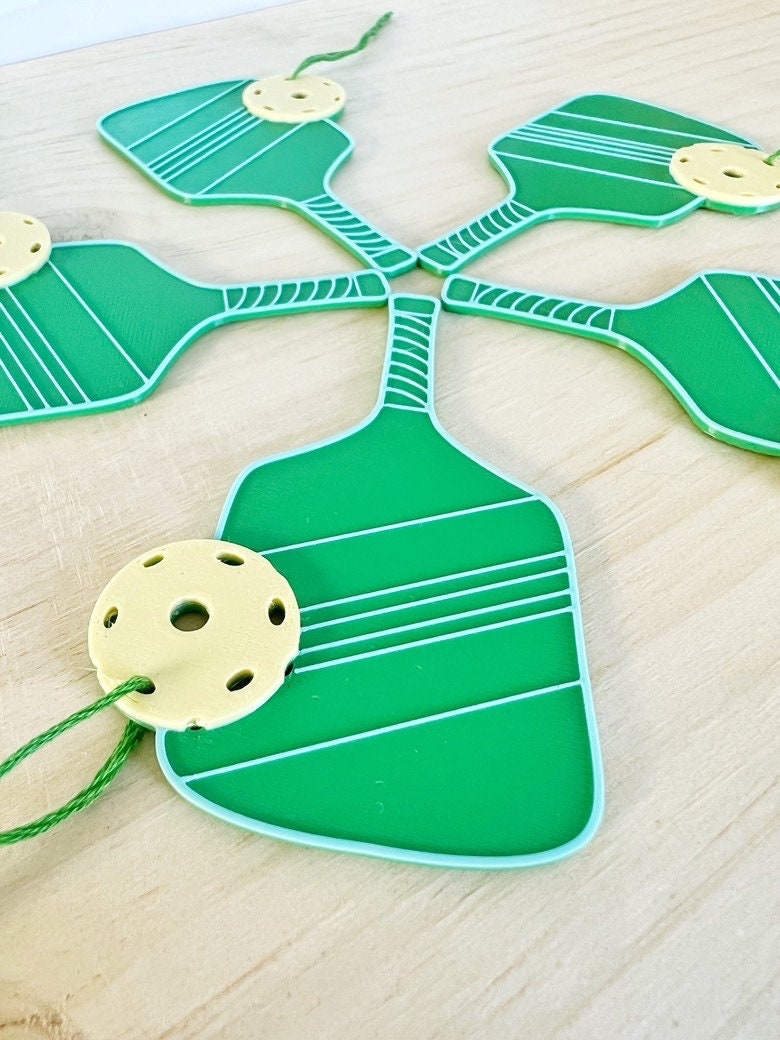 Close-up of a green 3D printed pickleball paddle ornament with a yellow ball, with more ornaments behind