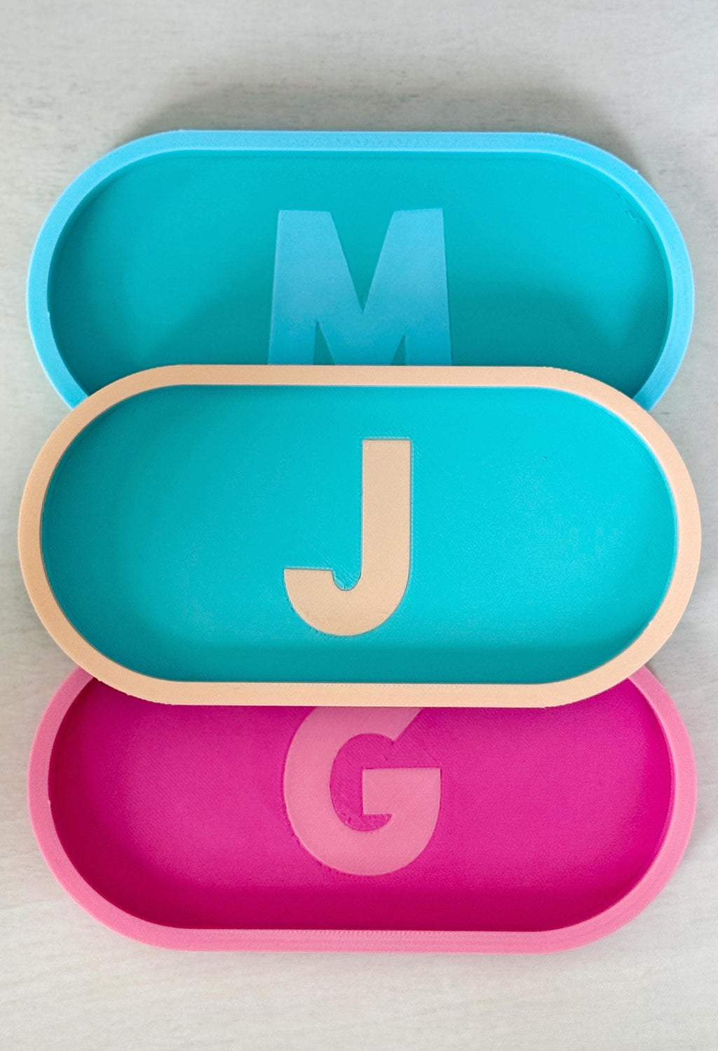 Stacked personalized initial trays with letters M, J, and G in teal, pink, and two-tone colors