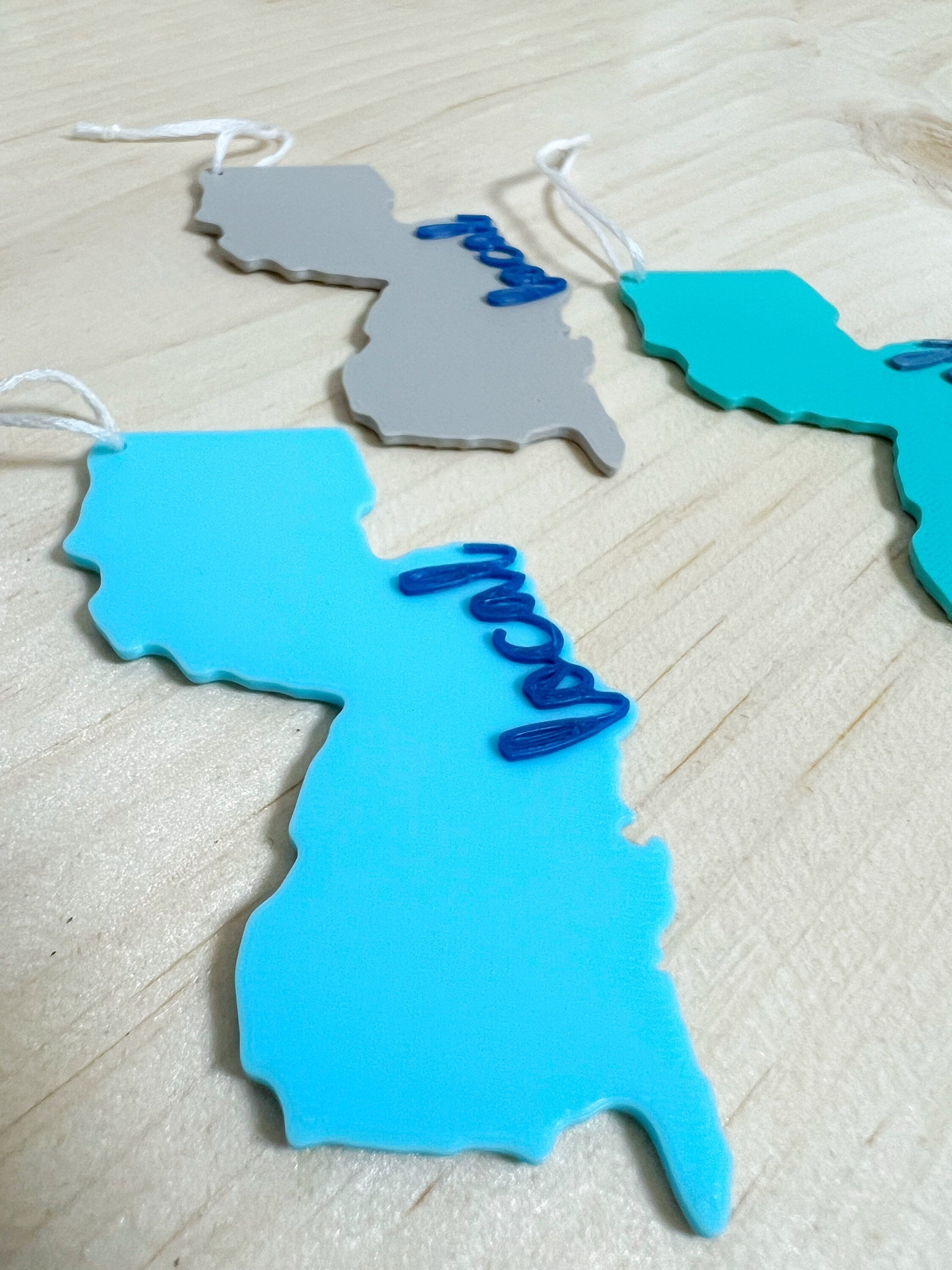 Close-up of a blue 3D printed New Jersey ornament with local in navy script, with gray and teal versions behind