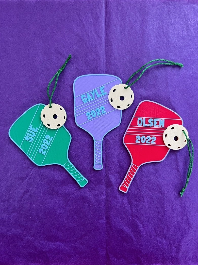 Three personalized pickleball paddle ornaments reading Sue 2022, Gayle 2022, and Olsen 2022 in green, purple, and red