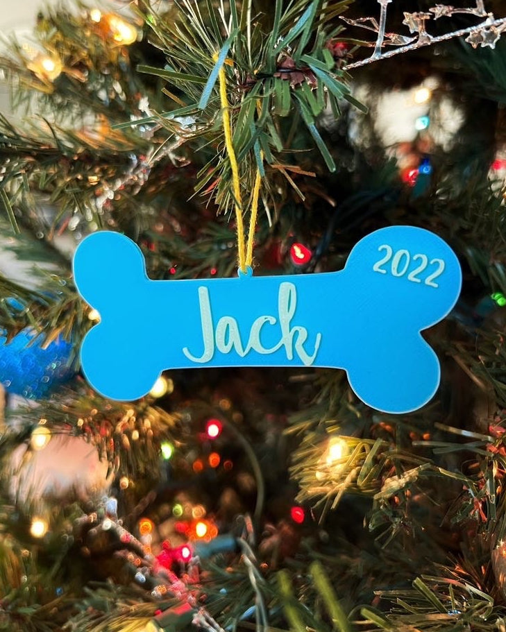 Personalized 3D printed dog bone ornament reading Jack 2022, light blue with mint lettering, hanging on a lit Christmas tree