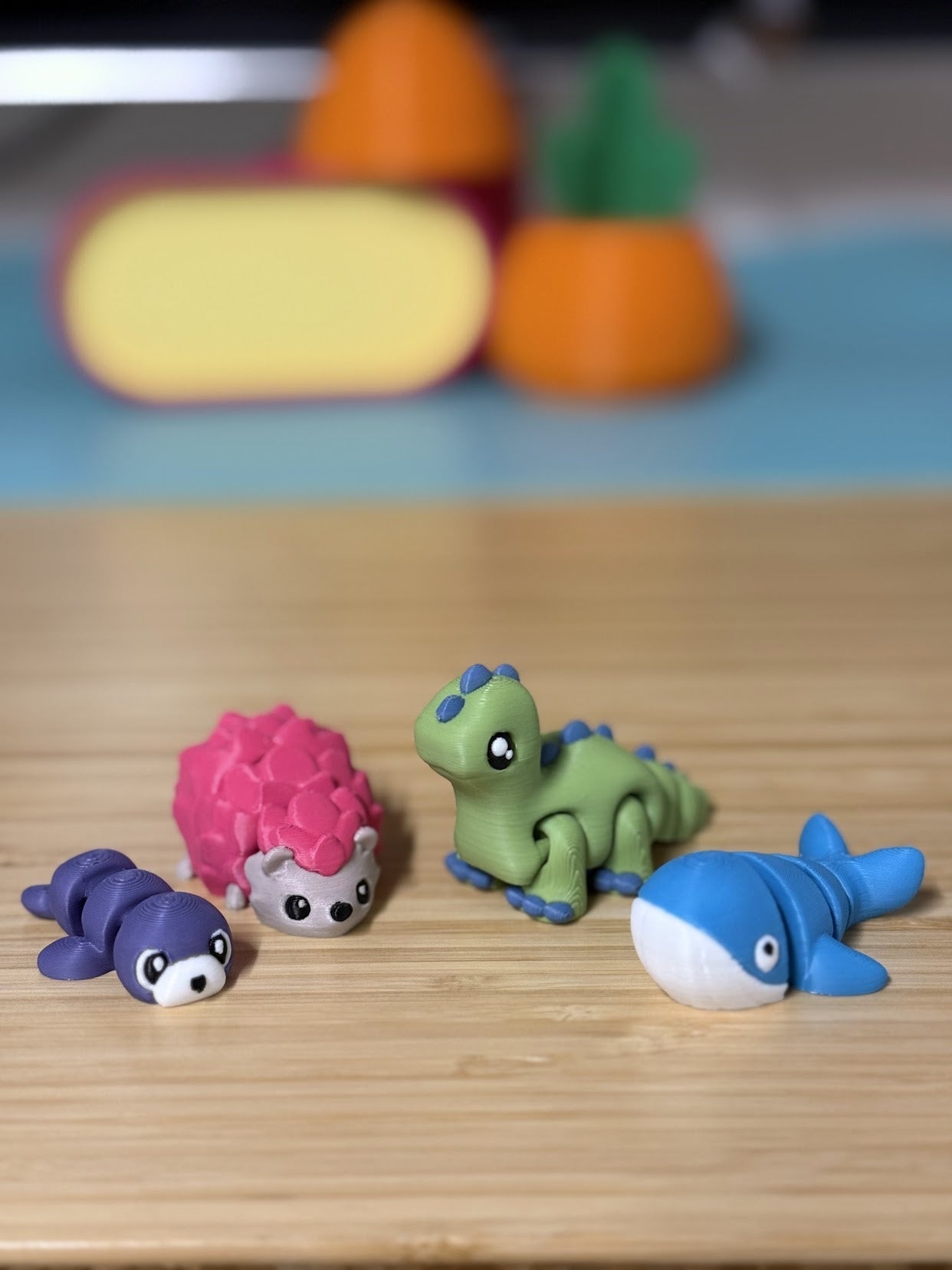 Green 3D printed Mini Flexi Brontosaurus with blue spikes next to a purple seal, pink hedgehog and blue whale flexi on a wood desk